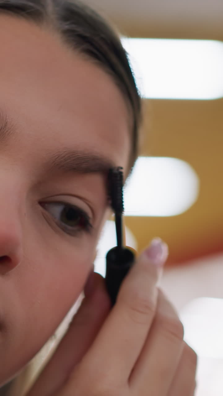 Adolescente blanco aplicándose máscara de pestañas en primer plano, detalle intenso en cejas y pestañas con mano firme guiando el pincel, luces de estudio y reflejos en el espejo, preparación tranquila y centrada, modelo practicando la técnica