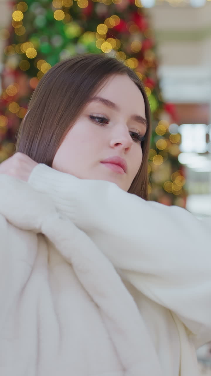 Woman putting on winter jacket in stylish shopping mall decorated with festive Christmas tree and warm lights, other shoppers are seated in the background