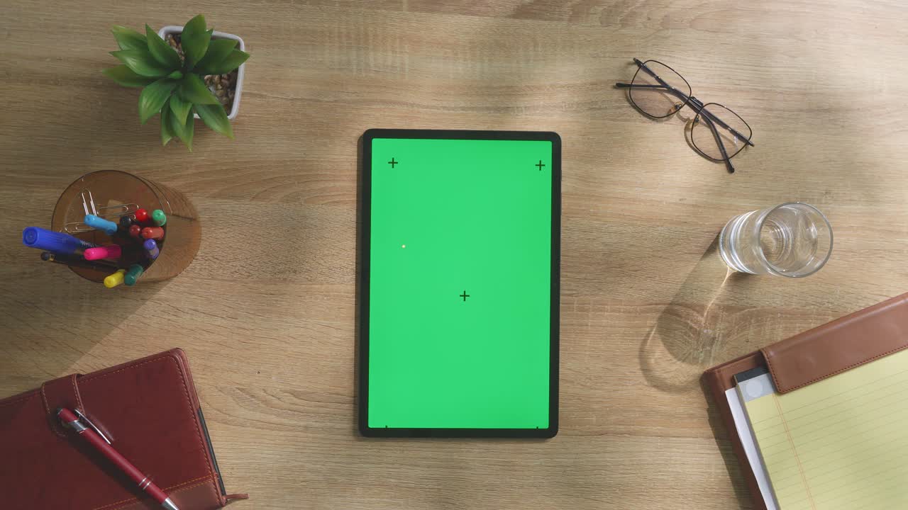 vista de arriba hacia abajo de una computadora portátil con pantalla verde simulada pantalla chromakey en un escritorio de oficina de madera al lado de la libreta con bolígrafos, gafas y un vaso de agua.