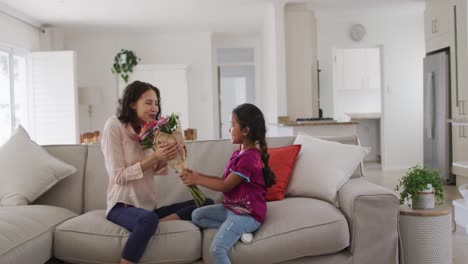 Happy-hispanic-woman-sitting-on-sofa-getting-flowers-from-daughter