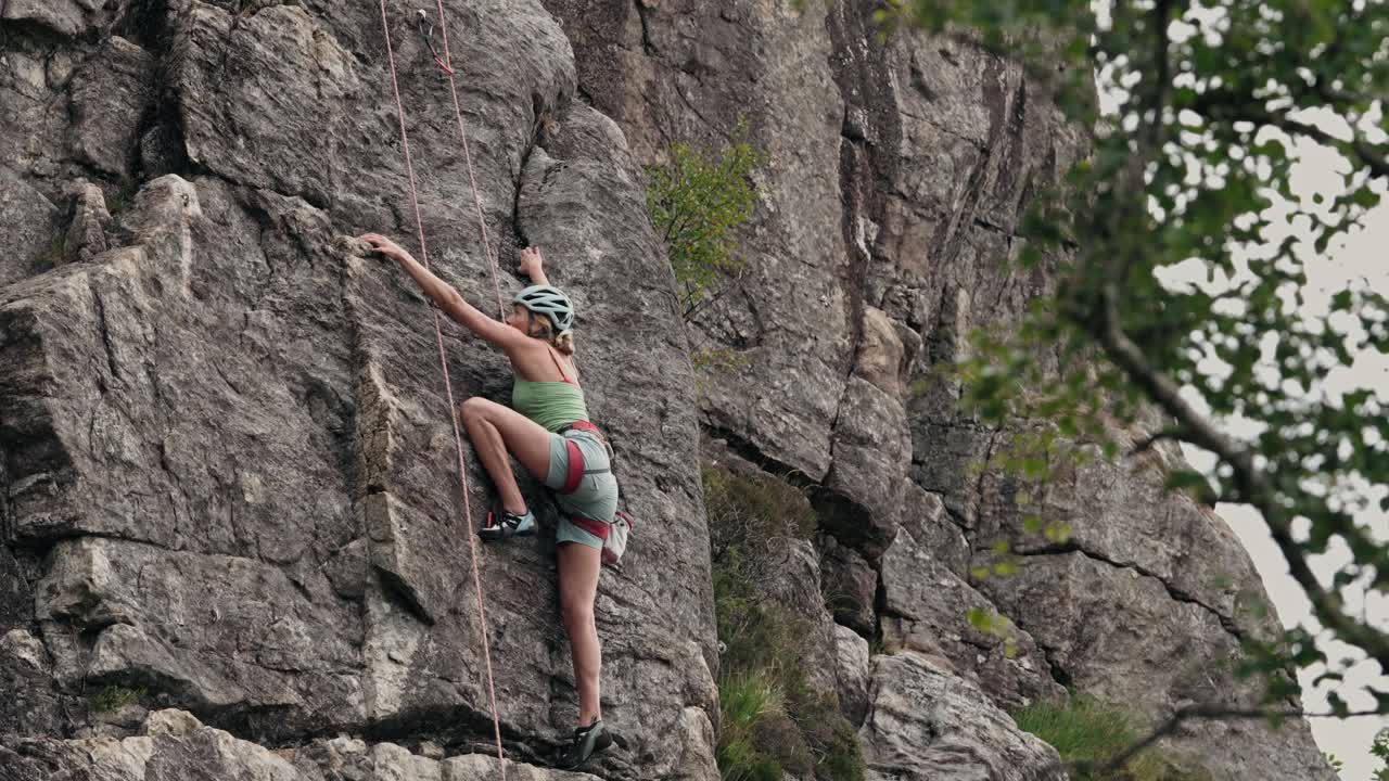 mujer joven con casco y cuerdas subiendo acantilado, vista lateral