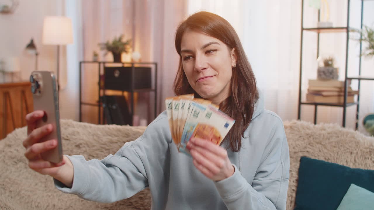 улыбающаяся женщина-блогер-инфлюенсер записывает историю на смартфоне, сидя на диване и показывая деньги дома.
