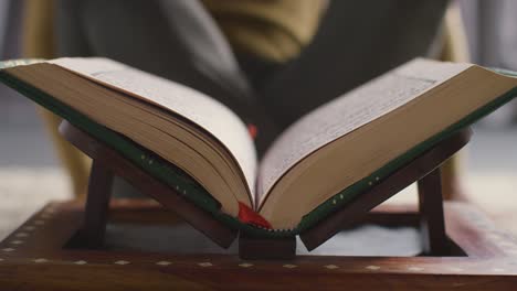 close up of open copy of the quran on stand at home with man sitting behind