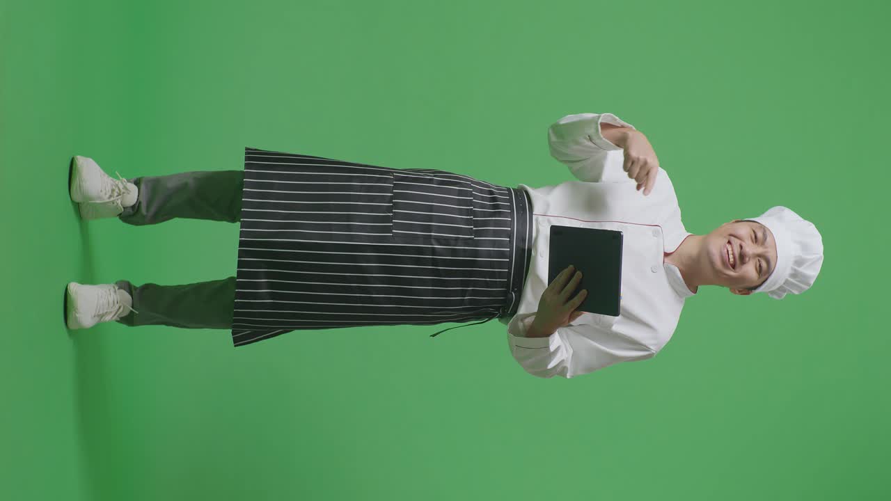 Full Body Of Asian Man Chef Smiling And Pointing To Tablet In His Hand While Standing In The Green Screen Background Studio
