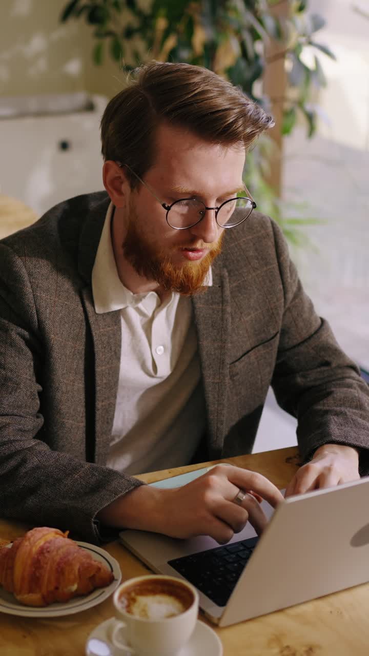 Giovane uomo che lavora al laptop in un caffè con caffè e croissant