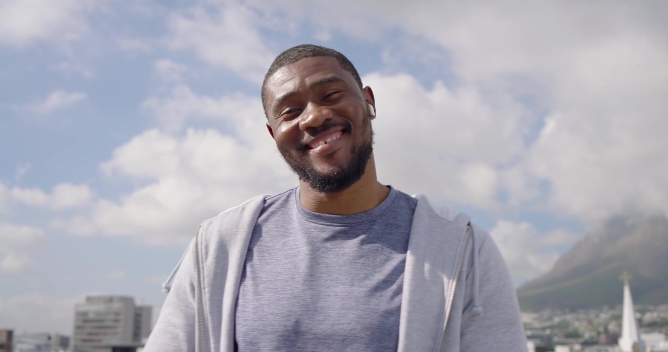 Mid-adult African man wearing earbuds and smartwatch, listening to music, rubbing hands on rooftop