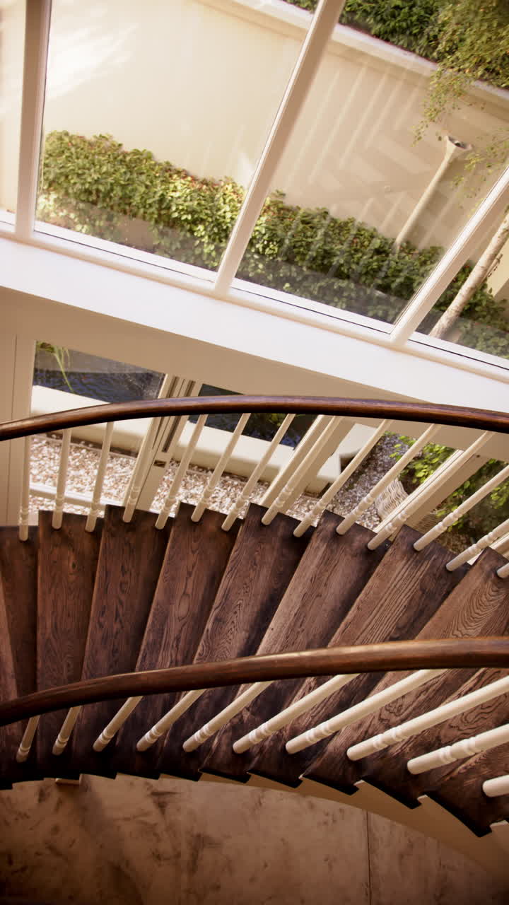 Vertical video: Curved wooden staircase with white railings leading to large windows with garden vie