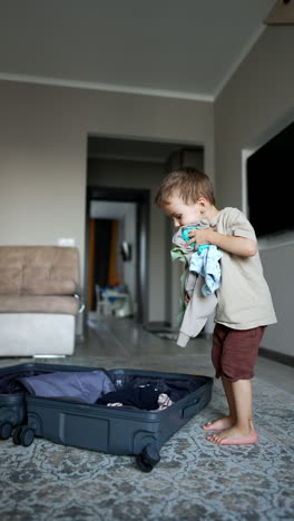 four-year-old toddler boy picks clothes from the suitcase. funny kid throws a pile of things up happily. vertical video.