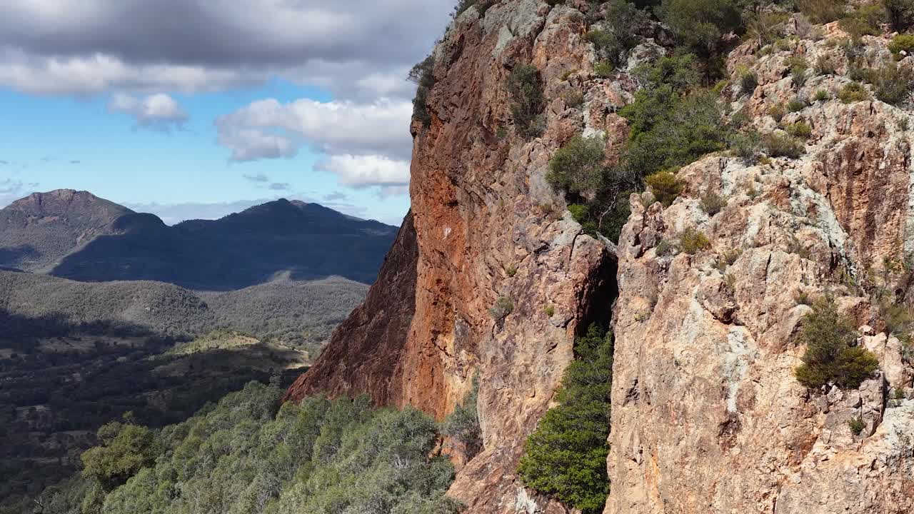 Camera slowly pans across Split Rock’s rugged cliffs, revealing distant forested hills under bright daylight with scattered clouds in Warrumbungle National Park