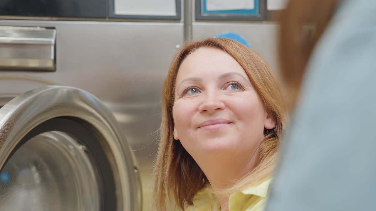 Mother smiles while teaching daughter cleaning at laundromat, wiping stainless washer door with blue cloth, giving guidance, building hygiene habit, family teamwork during chore, learning moment