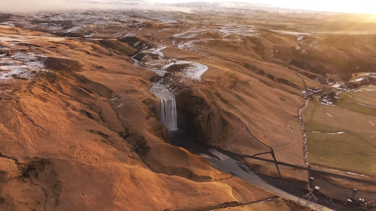 Golden hour aerial view of Skógafoss waterfall in southern Iceland, flowing from the Skógá River near the base of Eyjafjallajökull volcano, surrounded by rugged, snow-dusted terrain.