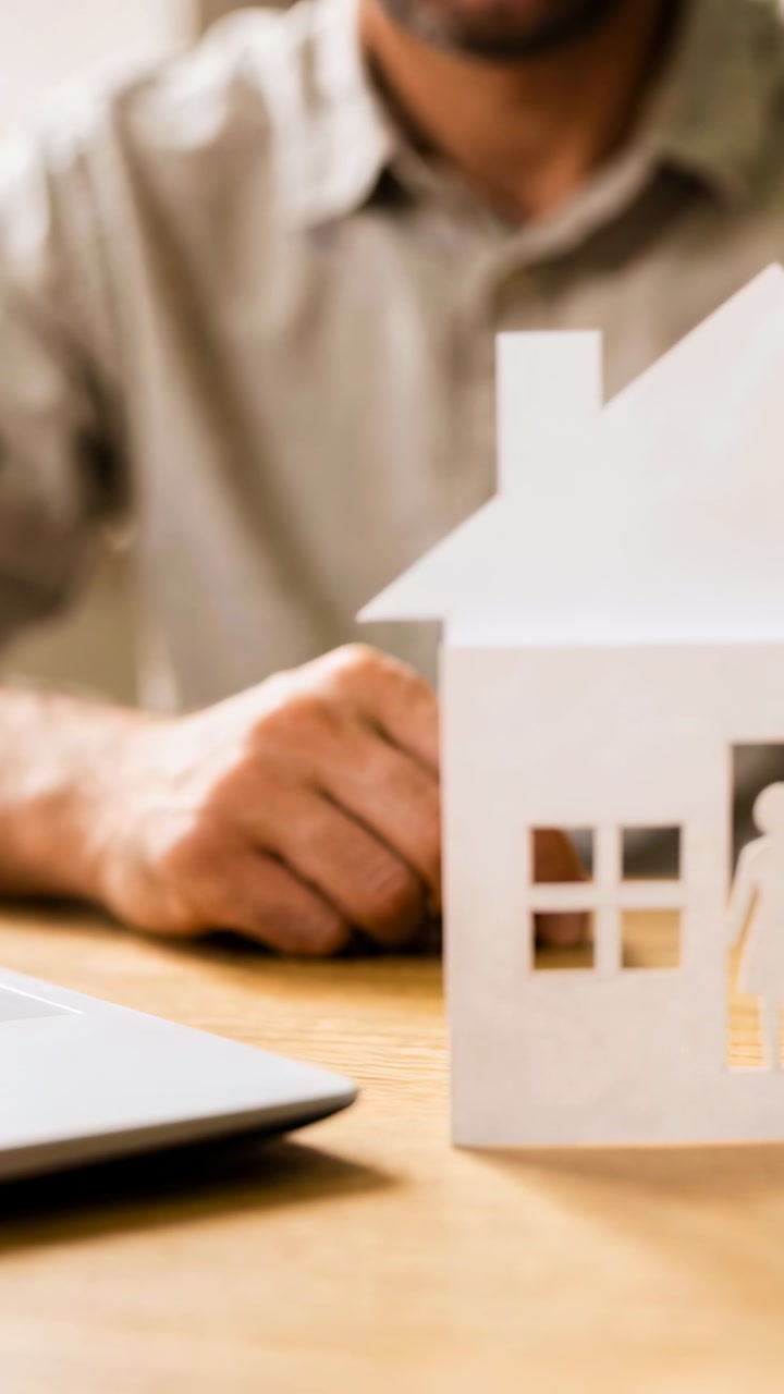 Vertical video: Moving man in shirt placing figure into paper house at desk, laptop, copy space