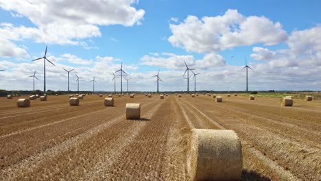 Die-Luftvideoaufnahmen-Zeigen-Die-Rhythmische-Bewegung-Von-Windturbinen-Vor-Dem-Frisch-Abgeernteten-Feld-Eines-Bauern-Aus-Lincolnshire,-Das-Mit-Goldenen-Heuballen-Geschmückt-Ist