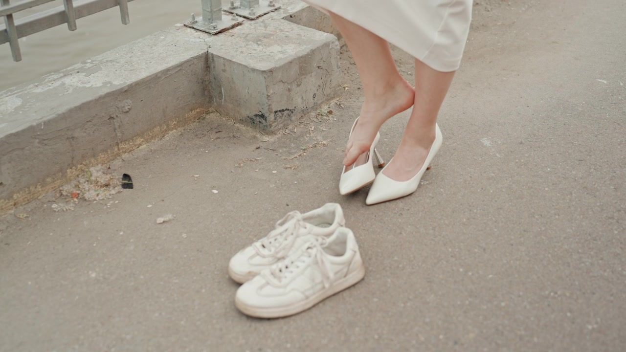 Mulher de vestido branco tira seus sapatos de salto alto pontudos e calça tênis casuais enquanto está em pé ao ar livre perto de estruturas de concreto. A cena captura a transição do formal para o confortável