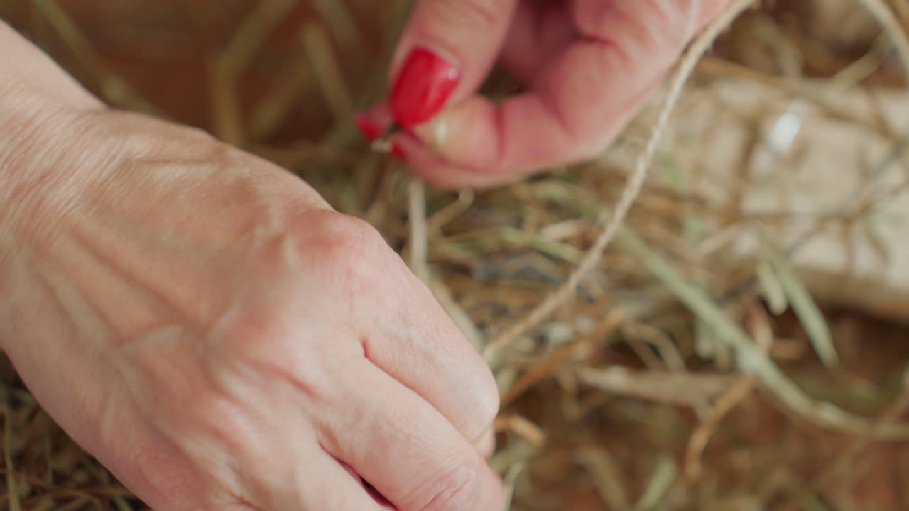 Decorator tying twine around dried hay with careful hands, securing natural material for rustic wreath craft, close-up view showing detailed process of handmade seasonal decoration in creative workspace