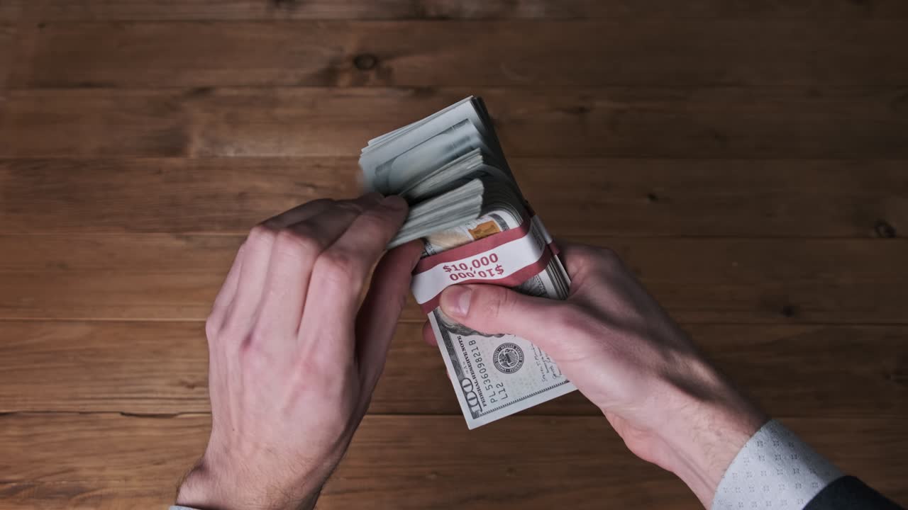 las manos masculinas sostienen tres pilas de billetes de 10.000 dólares en un fondo de madera.