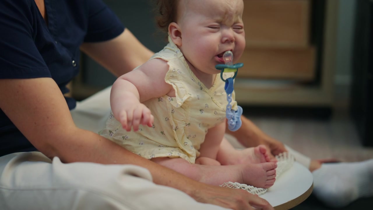 Baby sitting with adult support on a soft indoor mat, wearing a light yellow outfit during a calm caregiving moment