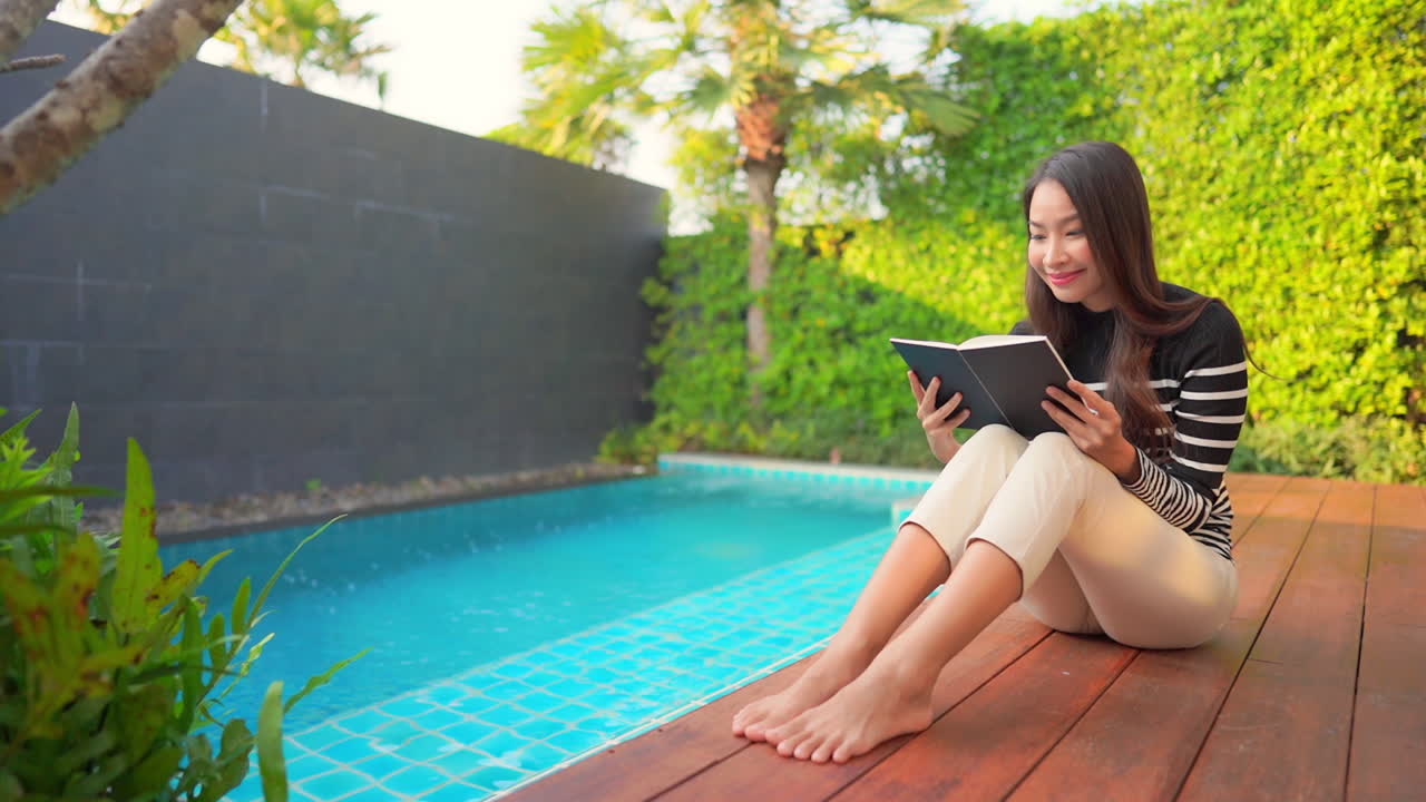 Beautiful asian woman relaxing on poolside smiling and reading a book outside