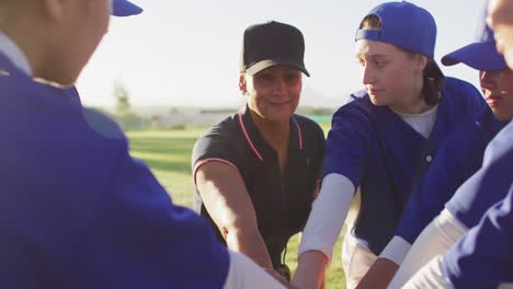 Diverse-group-of-female-baseball-players-and-coach-on-pitch,-in-a-huddle,-stacking-hands