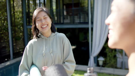 Happy-asian-female-friends-with-exercise-mats-and-water-bottle-laughing-on-terrace,-slow-motion