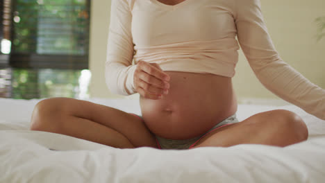 midsection of caucasian pregnant woman sitting on bed and putting headphones on belly