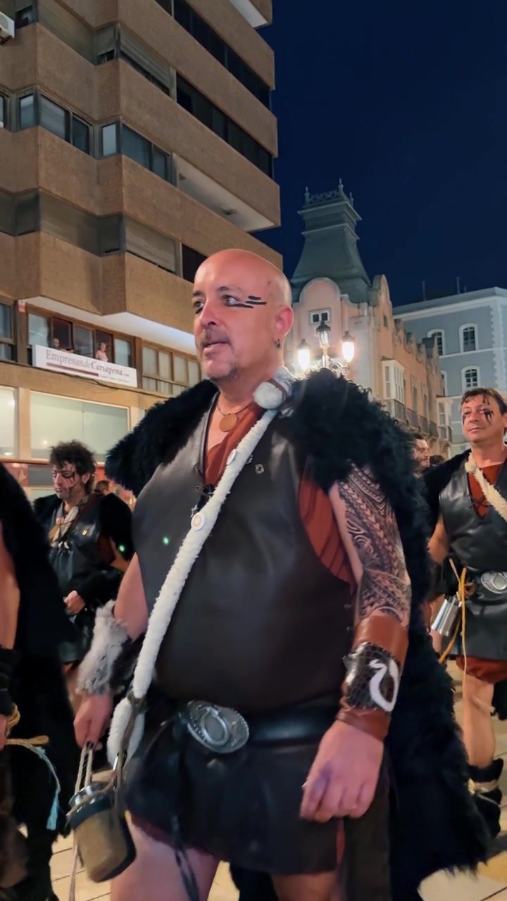 Alicante, Spagna, 8 agosto 2025: Uomini in costumi da barbari sfilano per le strade della città di notte. Processione di rievocatori che indossano gilet di pelle e pelliccia sintetica durante un festival notturno.