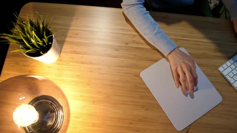 female working on computer at night. crop view of hands of woman in comfortable home clothes sitting at wooden desk lit by lamp and using computer mouse and keyboard in dark room at night.