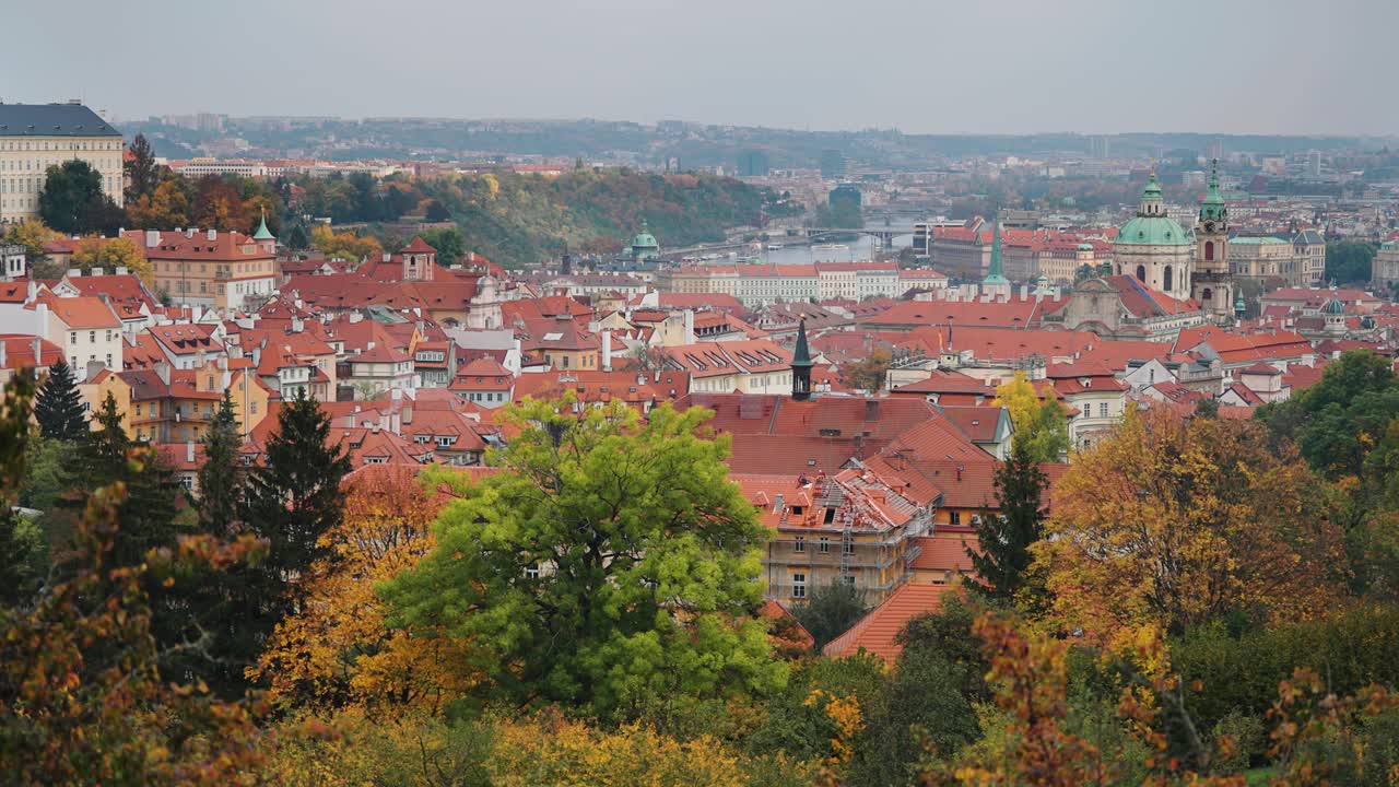 Una vista panoramica dei tetti rossi storici e delle cupole verdi di Praga, vista dalla collina di Petřín con alberi autunnali in primo piano.