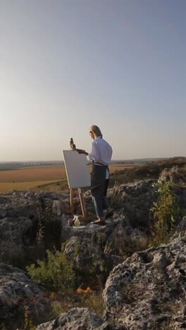 painting a landscape with sky water and buildings on canvas atop a rocky hill