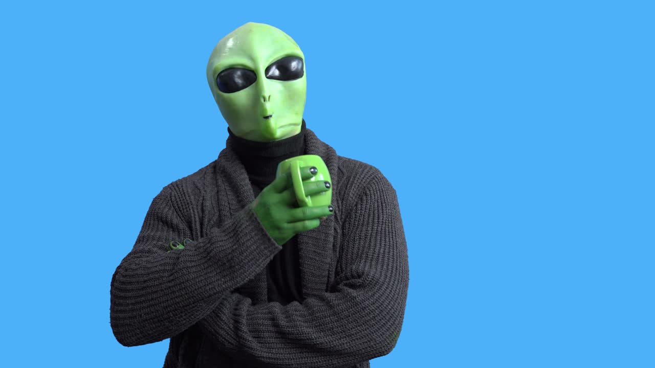 Professional actor in alien costume and cozy home outfit comes into the frame with funny mug and sits down to drink hot beverage against blue chromakey background