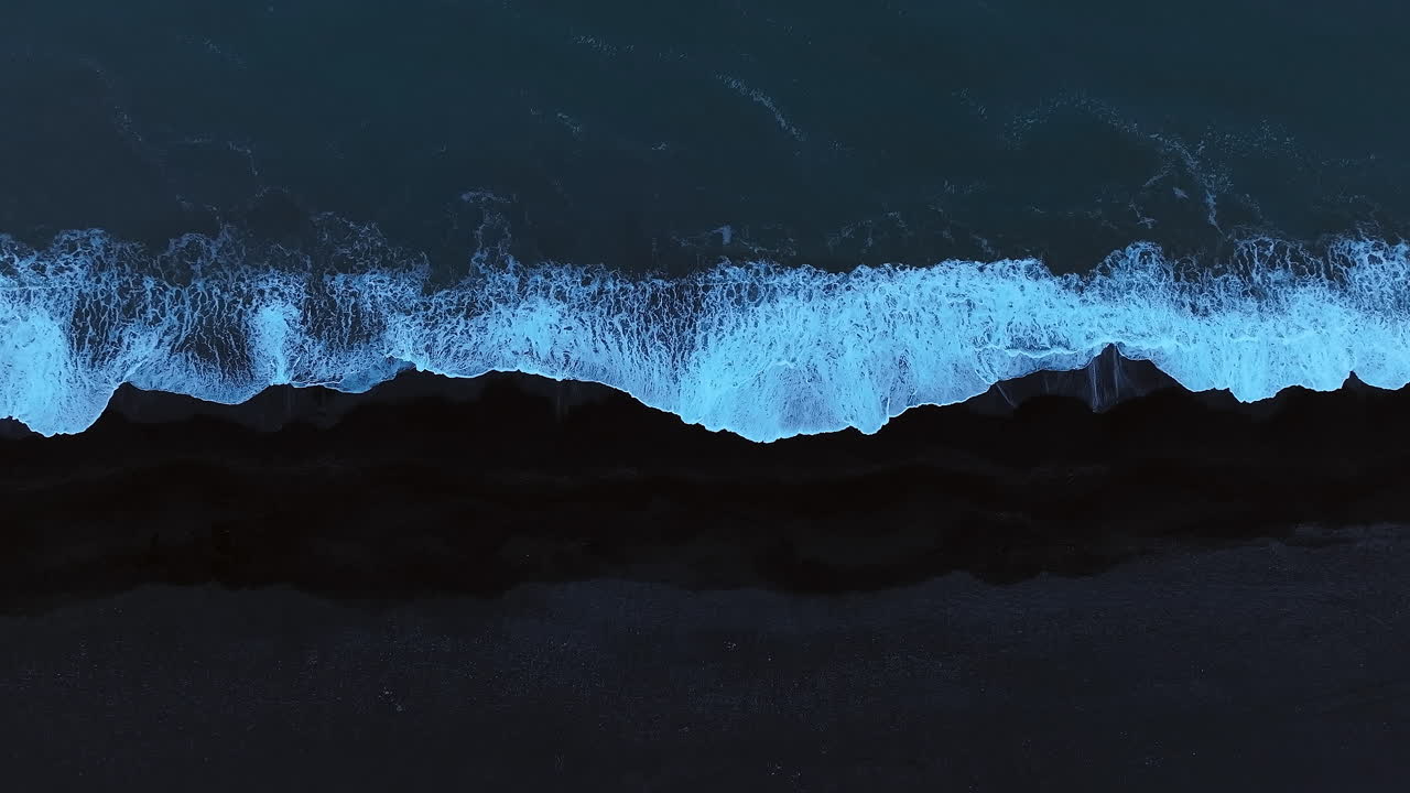 Dark blue ocean waves splashing black sand beach, aerial top down shot