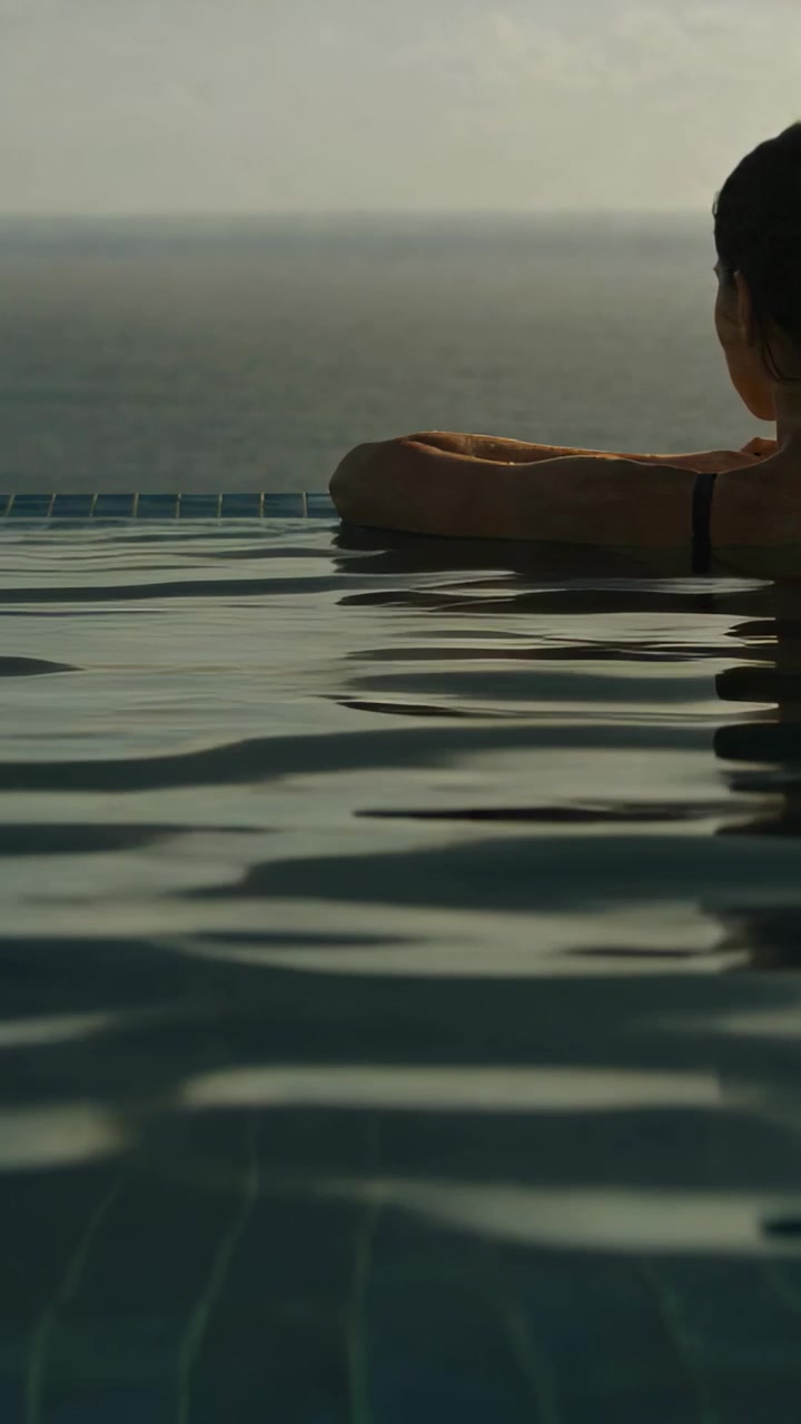Vertikales Video: Sich lehnende Frau blickt vom Rand eines Infinity-Pools aufs Meer, Badeanzugträger, Platz für Text.
