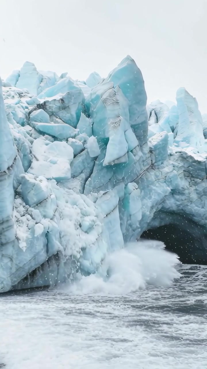 Video verticale: Fronte di ghiacciaio blu-bianco che si scioglie, rilasciando una lastra di ghiaccio nel mare in una grotta marina, creando schiuma