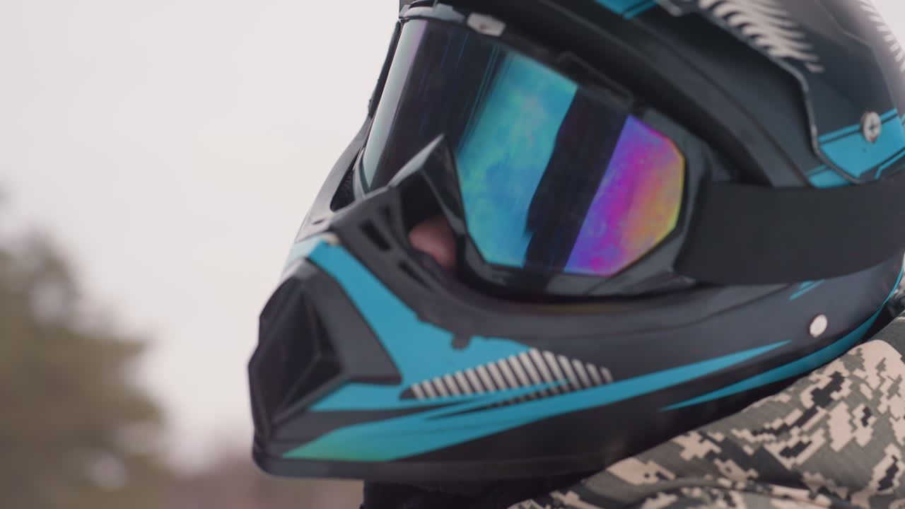 Helmeted rider wearing mirrored goggles, closeup profile of motocross athlete in teal and black full face helmet and camouflage jacket, iridescent lens reflecting snowy overcast forest, poised