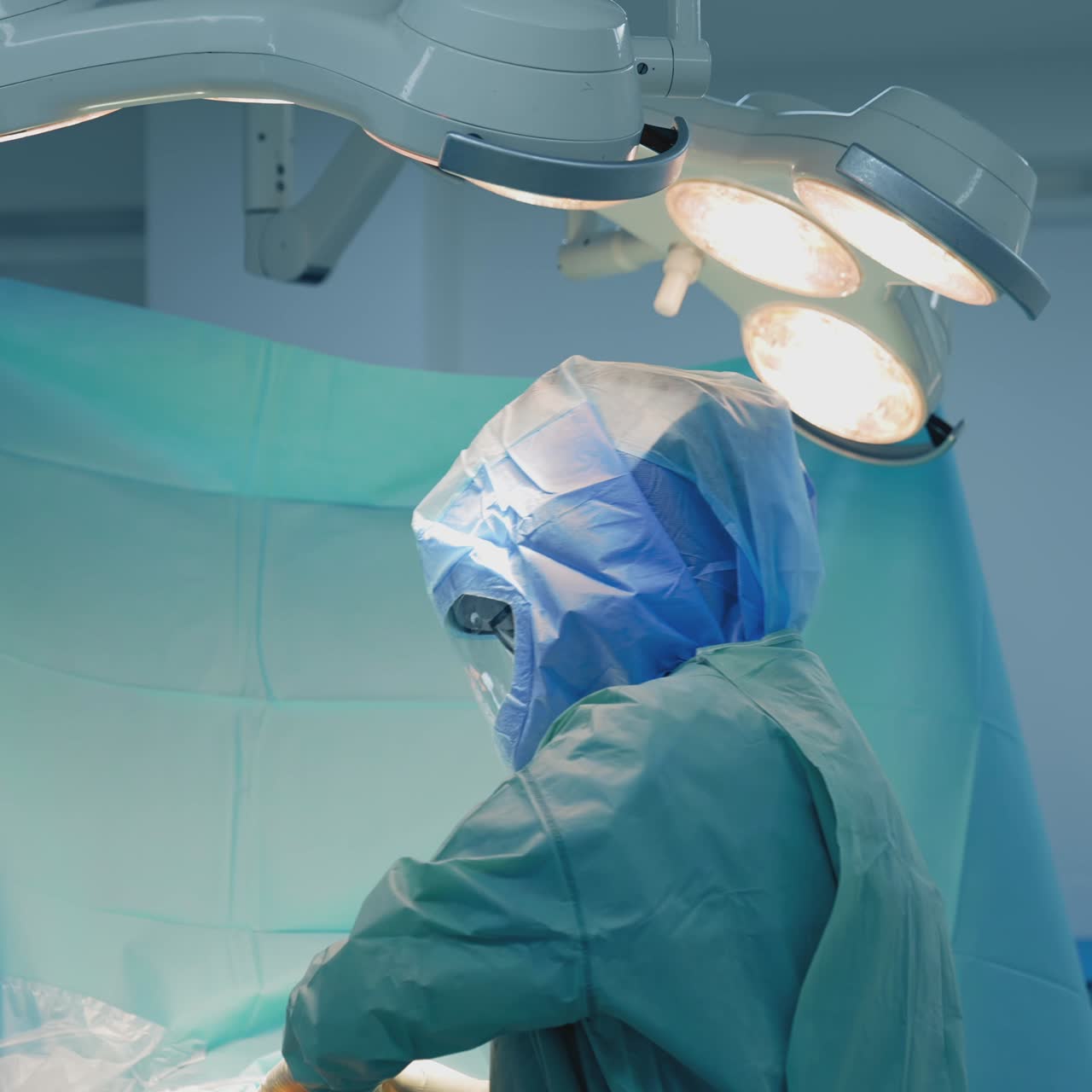 Doctor in protective uniform suit in clinic. Medical specialist in safety helmet performing a surgery under the surgical lamp in the operating room. Coronavirus pandemic