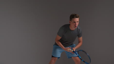 studio shot of male tennis player returning shot in match hitting ball with racket against grey background