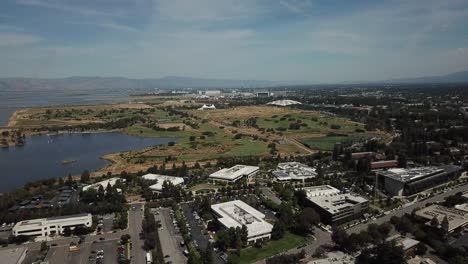 Aerial-birds-eye-view-of-shoreline-lake-Google-campus-amphitheater-park-mountain-background-move-forward