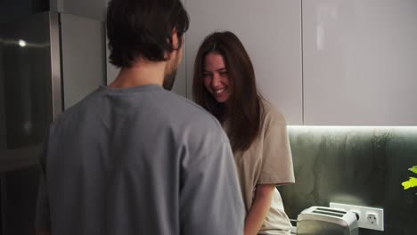 Close-up-view-over-the-shoulder-of-a-happy-brunette-guy-giving-a-gift-in-the-shape-of-a-red-heart-to-his-brunette-girlfriend-in-a-beige-T-shirt-in-a-modern-apartment-in-the-evening