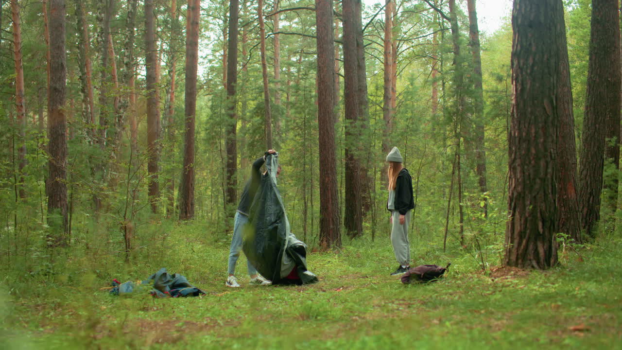 Young man prepares to set up tent while girl stands nearby observing in serene forest environment surrounded by tall trees and green vegetation, backpacks and camping gear scattered on grass