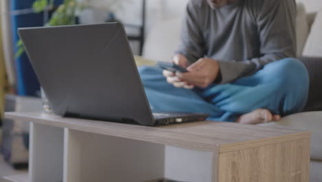 Pijamas-Trabajando-Desde-Casa,-Un-Joven-Adulto-Usa-La-Sala-De-Estar-Como-Oficina-Junto-Con-Una-Computadora-Portátil,-Se-Distrae-Con-Una-Ventana-Emergente-Móvil-Sobre-Un-Nuevo-Mensaje