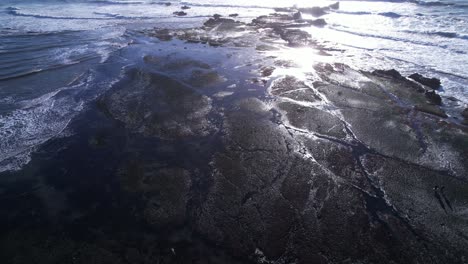 drone jib up and tilt down over surfers, tide pools and crashing waves at mavericks beach, california during sunset