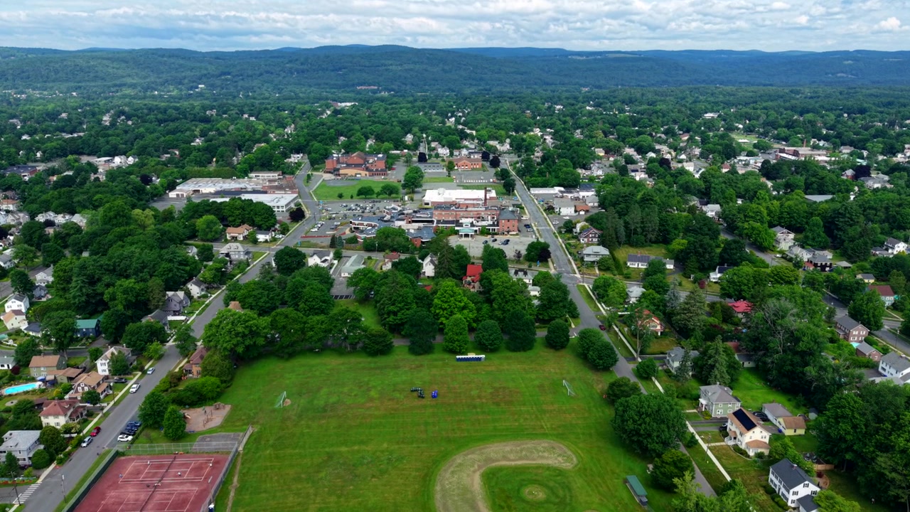 Drone in lontananza vola verso il Baystate Franklin Medical Center in un quartiere residenziale con area verde e campo sportivo circondato da alberi, Greenfield, Massachusetts, USA