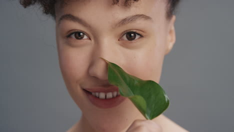 close up portrait beautiful mixed race woman smiling caressing face with leaf touching smooth healthy skin complexion enjoying natural skincare beauty on grey background