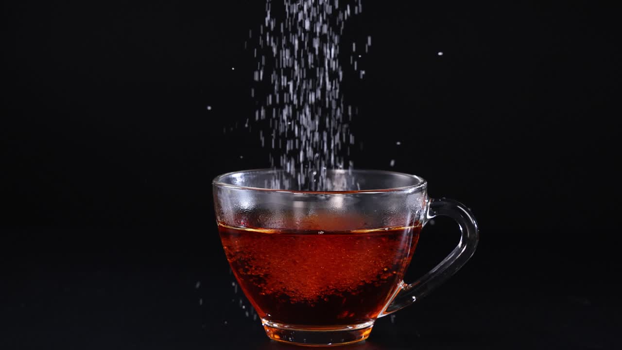 Sugar granules cascade into a glass cup of tea against a dark backdrop, creating a dynamic visual effect