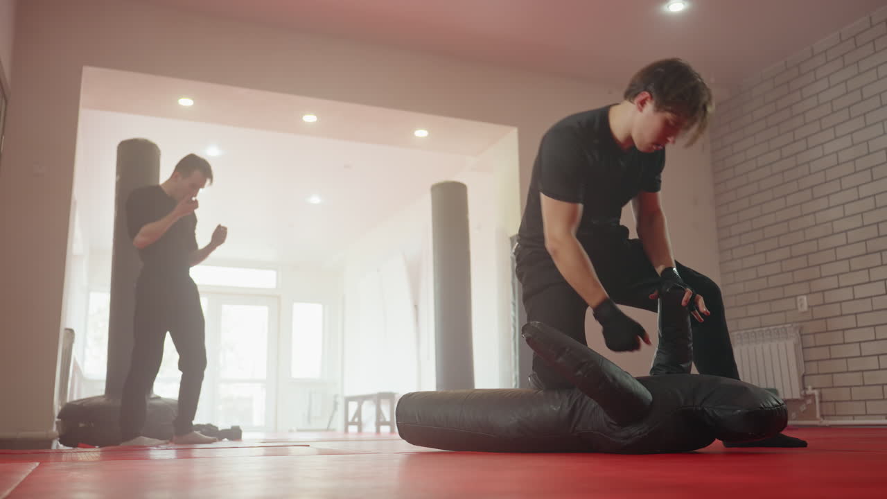 Policeman in black uniform practicing ground combat training on dummy inside gym while another man observes from background, showcasing discipline, strength, resilience during intensive martial arts