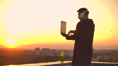 Un-Hombre-Independiente-Con-Auriculares-Parado-En-El-Techo-Al-Atardecer-Escribe-En-Las-Páginas-De-Códigos-Del-Teclado.-Pequeño-Negocio.-Escuche-Música-Y-Trabaje-En-La-Computadora-Disfrutando-De-La-Hermosa-Vista-Desde-La-Azotea.