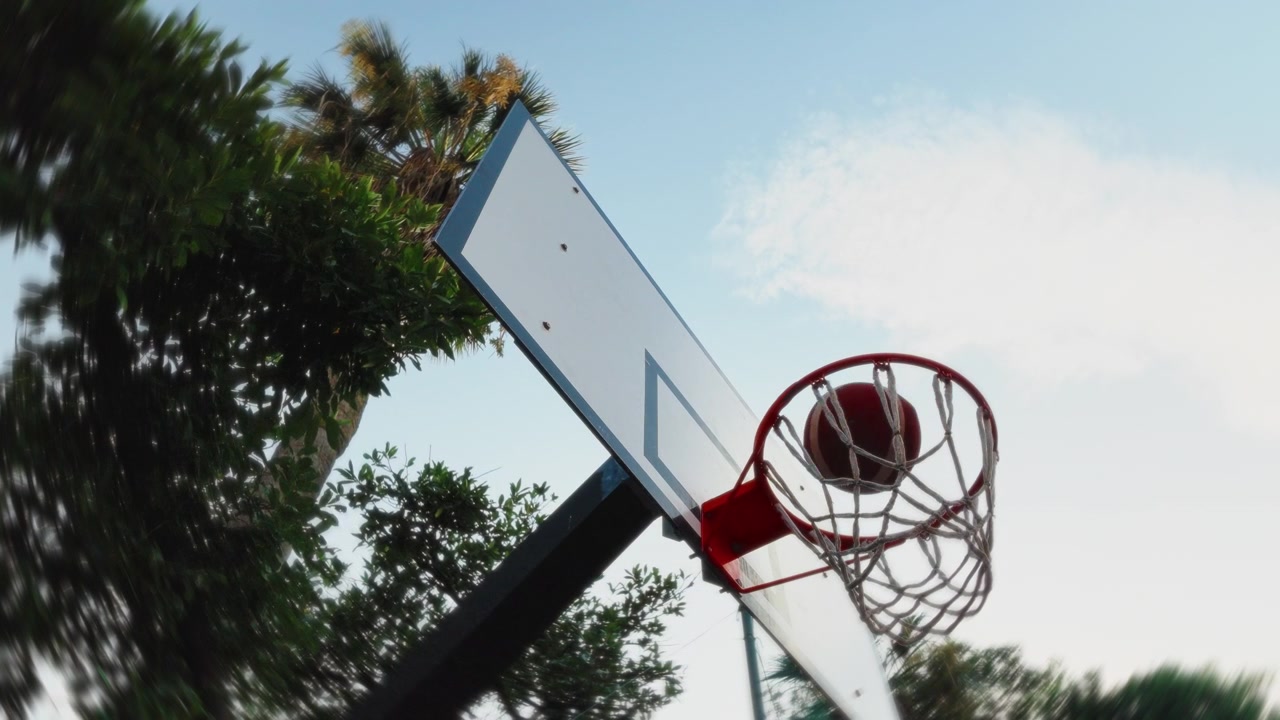 Una palla da basket cade attraverso un canestro esterno al rallentatore con alberi e cielo in vista