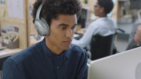 young mixed race businessman using computer working on business project browsing online listening to music brainstorming ideas wearing headphones in relaxed office workplace