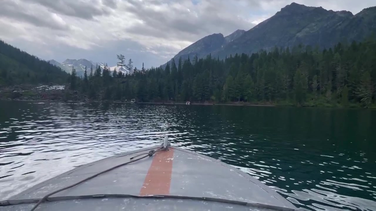 Forward view from a small boat crossing Multinsky Lakes in the Altai Republic, Russia, with dark conifer forest, rocky shore and high mountains under cloudy sky