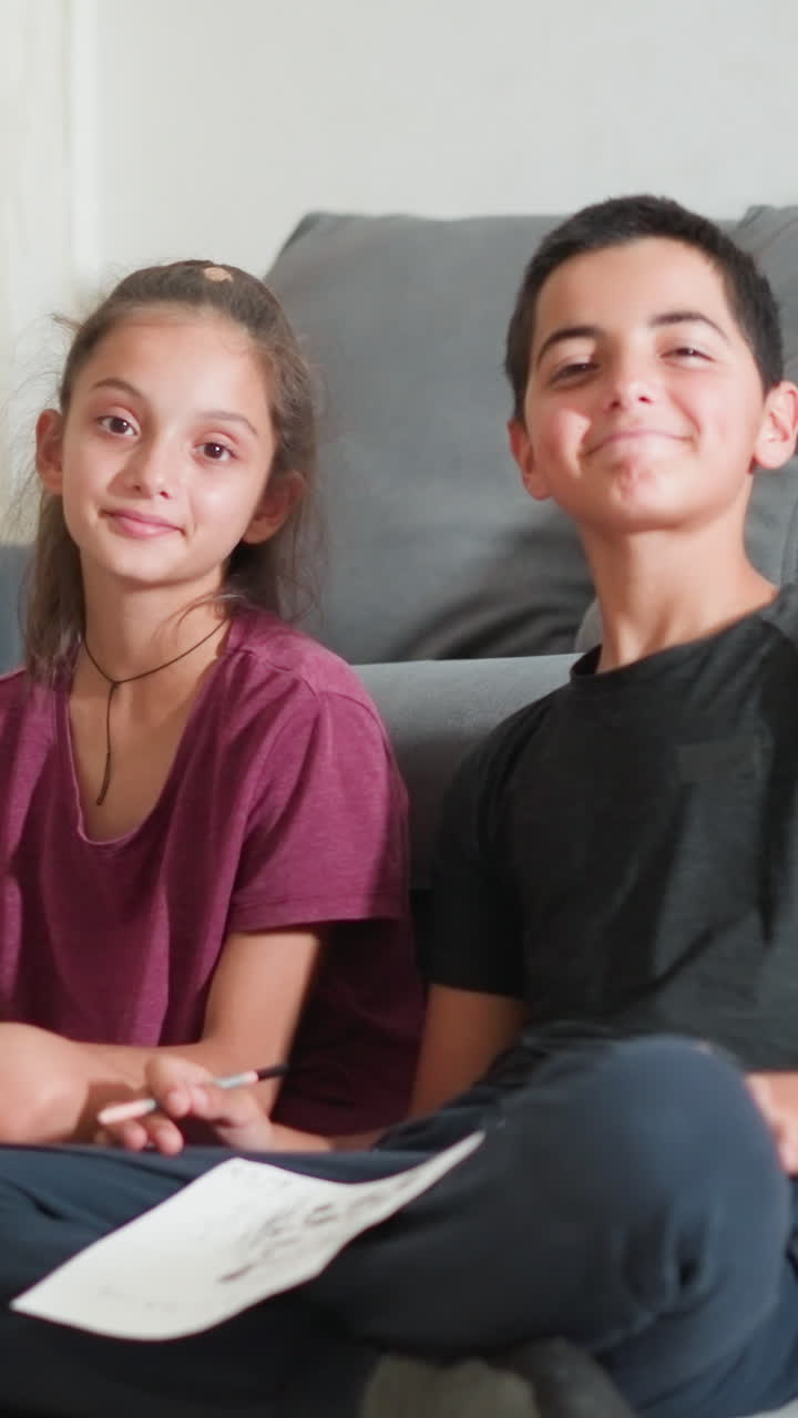 Primos adorables sentados en el suelo cerca del sofá, sujetando un papel y sonriendo con calidez a la cámara en un ambiente interior acogedor, mostrando la alegría de la actividad creativa compartida y el tiempo de unión con expresiones amistosas y relajadas